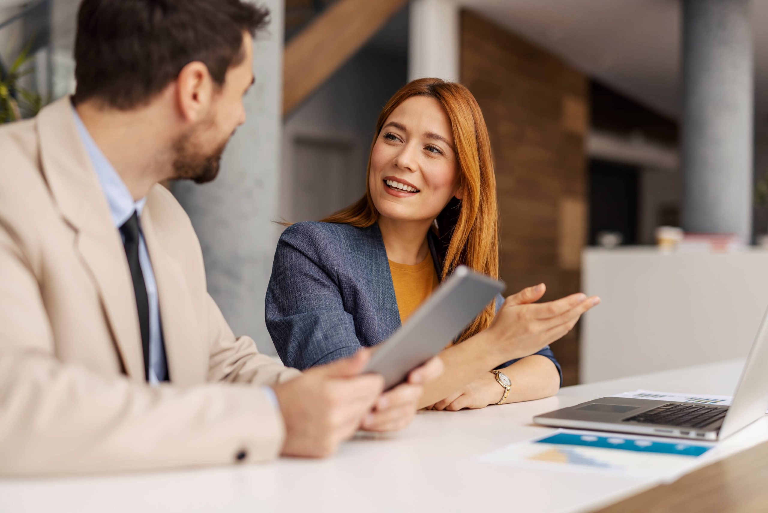 Businesswoman smiling and presenting to colleague with tablet and laptop on the desk